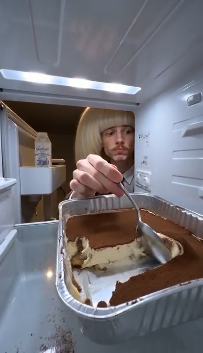 A person with a blond bob reaching into the fridge for a spoonful of tiramisu.