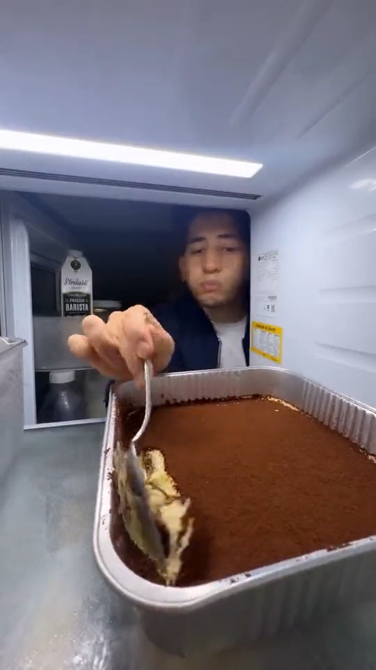 A man in a dark jacket holding a spoon in front of a tray of tiramisu.